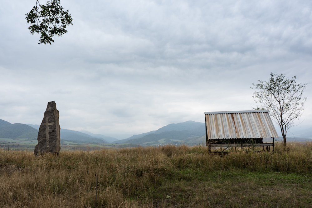 Una piedra con una cruz tallada en ella se alza junto a un refugio en desuso. Ambos se encuentran en lo alto de una colina en la frontera armenia, con las montañas de Azerbaiyán a lo lejos.