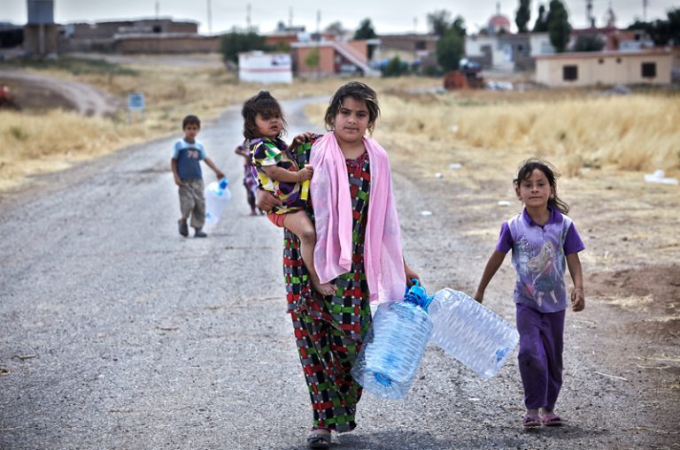 Una mujer iraquí de Mosul sostiene a su hija en uno de sus brazos mientras que con el otro acarrea un bidón de agua vacío. Sus otros dos hijos la siguen para ir a buscar agua cerca del campamento temporal de Garmawa. Fuente: ©ACNUR/UNHCR / S. Baldwin